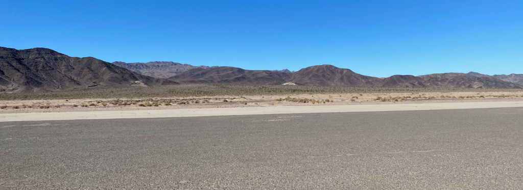Baker airport runway with gravel in the foreground and desert and mountains in the background with a clear blue sky.