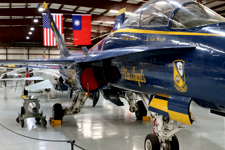 A Blue Angels plane is seen on the right-hand side of the photo parked inside a airport hangar museum with another WWII smaller plane in front of it under an American Flag and another flag that is red with a sun placed in the left of the flag.