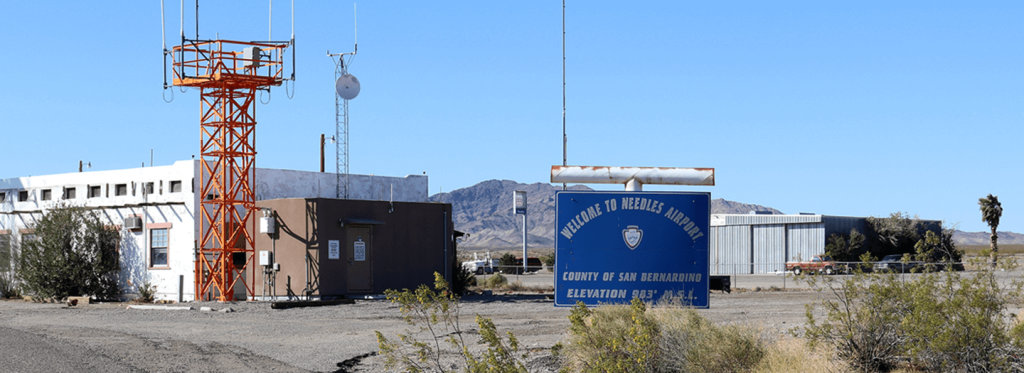 A welcome to Needles Airport sign is on the right and a building and tower with a platform is on the left with a satellite dish, mountains and grey metal hangar in the background.