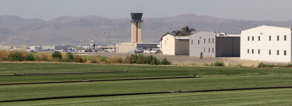The Chino Airport control tower is seen in the middle next to a helicopter in flight to the right facing the camera surrounded by agricultural fields and hangars on the right-hand side of the field.