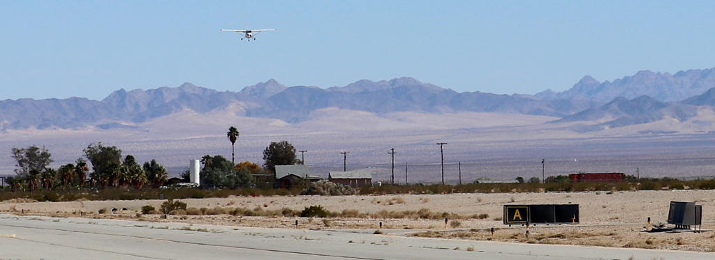 A small plane is seen coming in for a landing on the runway with house in the background and desert and mountains with a clear blue sky.