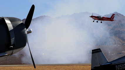 An airplane propeller is seen on the left while a vintage plane in flying over on the top right and a airplane tail is seen on the lower right. Smoke is seen in the foreground with desert mountains in the background.