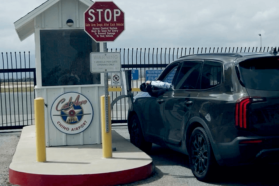 A vehicle is pulled up to the Chino Airport gate entrance with a man sticking his hand out the window to swipe a gate card to open the gate with a security office next to the gate access card system.