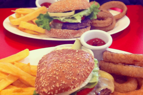 Two plates are shown across from each other with cheeseburgers, French fries and onion rigs on each plate with a little cup cup of ketchup.