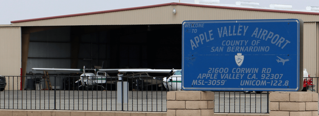 An airplane hangar is seen in the distance with a plane inside and a large sign is on the righ0hand foreground that says Apple Valley Airport San bernardino County 21600 Corwin RD Apple Valley CA 92307 MSL-3059 UNICOM-122.8