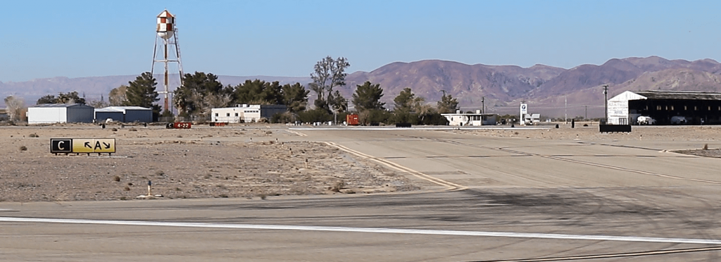 A tall water tower and buildings can be seen in the background against the mountains and airport runway signs can be seen in the foreground.