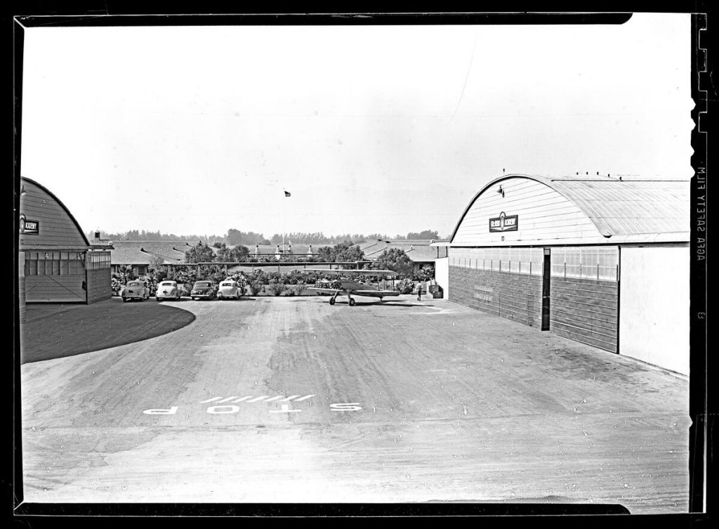 Photograph of an airplane outside hangars at the Cal-Aero Flight Academy, [s.d.], possibly World War II era (later, the Chino Airport). A small bi-plane can be seen at center, while hangars can be seen at left and at right. A sign reading, "Cal-Aero Flight Academy" can be seen above the doors on the hangars. A man can be seen standing to the left of the hangar at right, while four parked automobiles can be seen to the right of the left-most hangar. Bushes, an American flag, buildings, and an open field can be seen in the background. "Stop" can be seen painted upside-down on the concrete in the foreground.