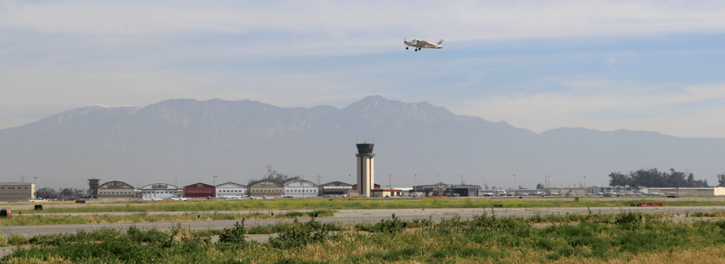 A control tower and hangars are seen in the background of the runay as a plane takes off and is in flight above the ground.