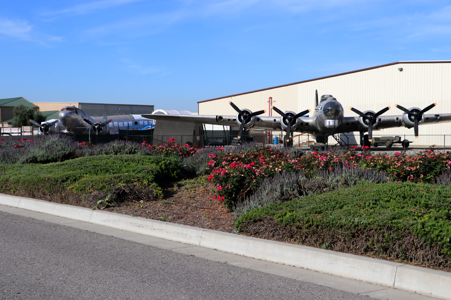 An exterior view of the Planes of Fame Air Museum. Center are silver WWII planes with the museum hangar building in the background surrounded by a chain link fence and colorful flowers and shrubs in the forefront in front of the paved street with a high cement curb.