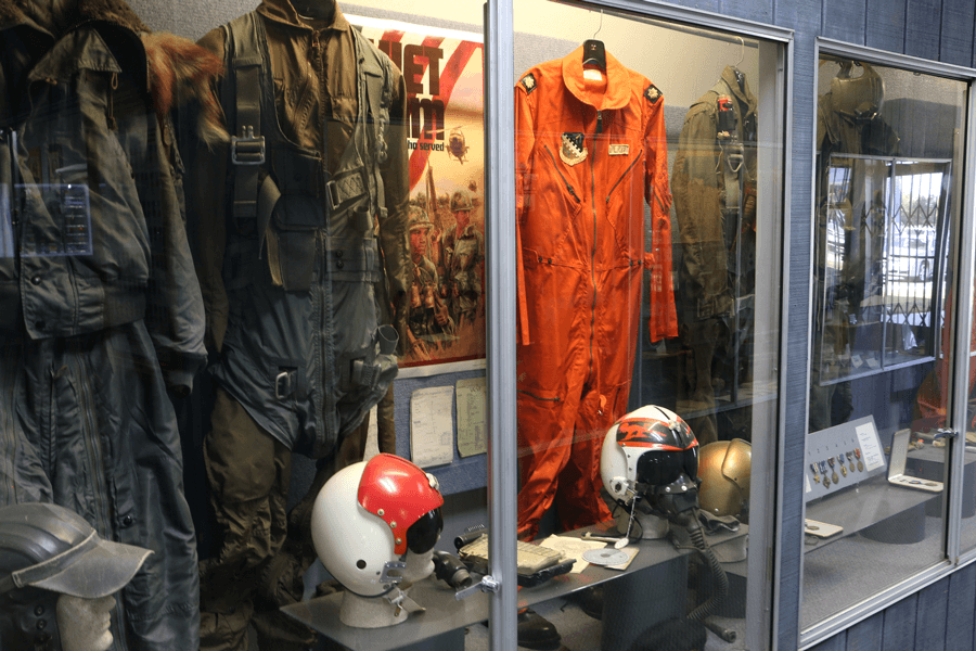 A trio of vintage flight suits are on display in a locked glass case with helmets.