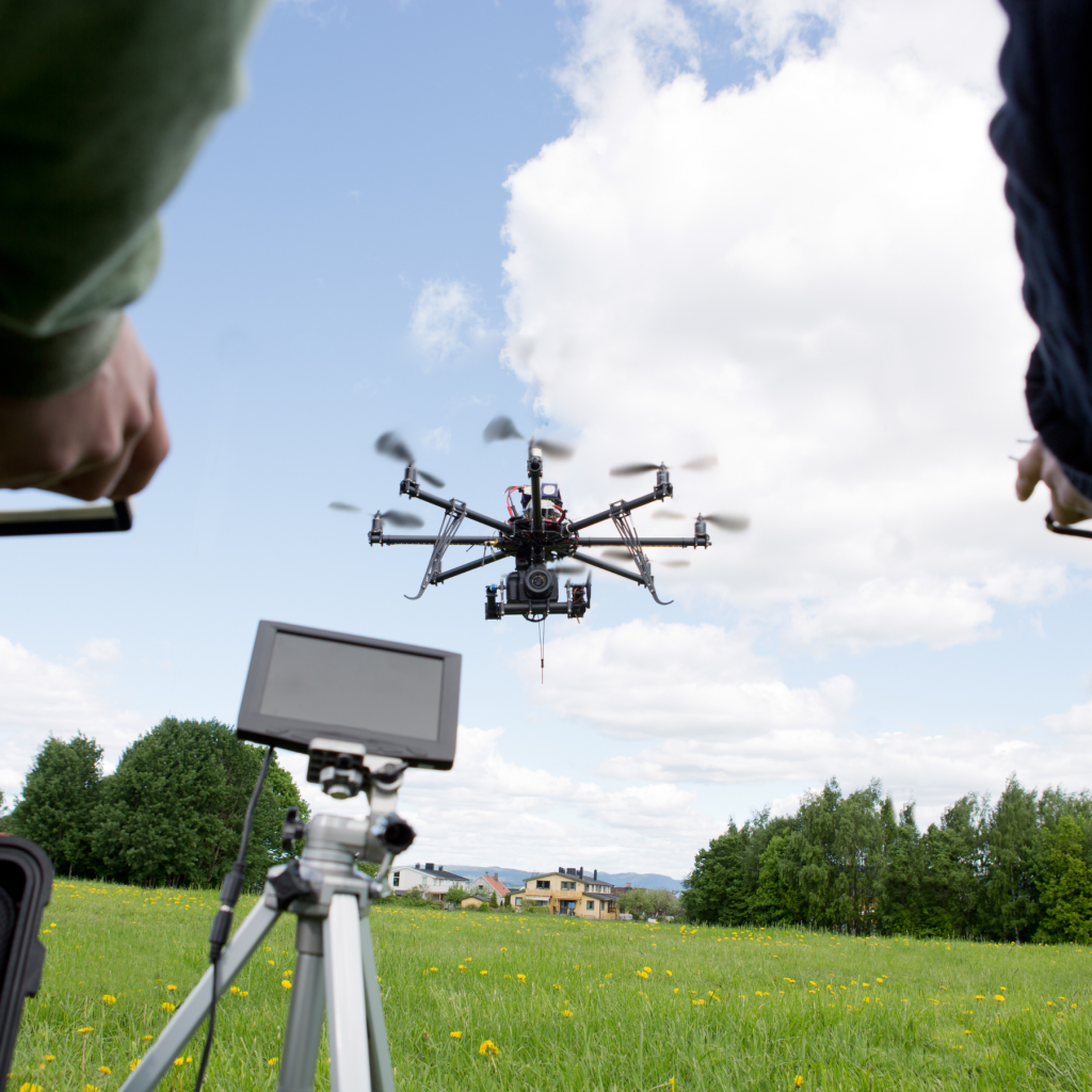 A drone is seen center in the sky in front of some white puffy clouds flanked by two people and a tripod in the middles with a screen on it.