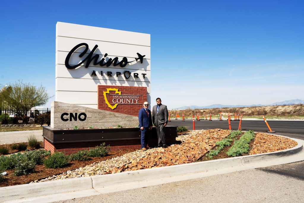 Two men in suits stand in front of a large sign for Chino Airport in San Bernardino County, California, on a clear, sunny day. The sign features the airport code "CNO" and is surrounded by landscaped rocks and plants, with traffic cones and a wide road in the background.